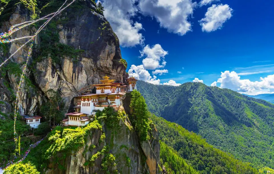Klasztor Paro Taktsang przyklejony do pionowej skały, wokół zielone góry i flagi modlitewne.