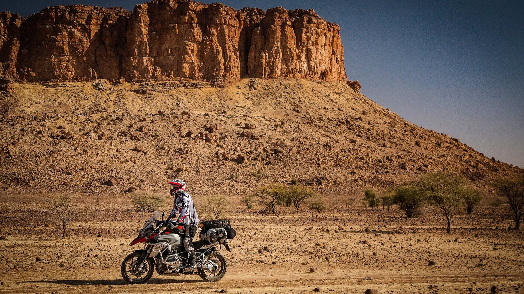 Motocykl z sakwami na pustynnym piachu, w tle rdzawe klify Sahary.